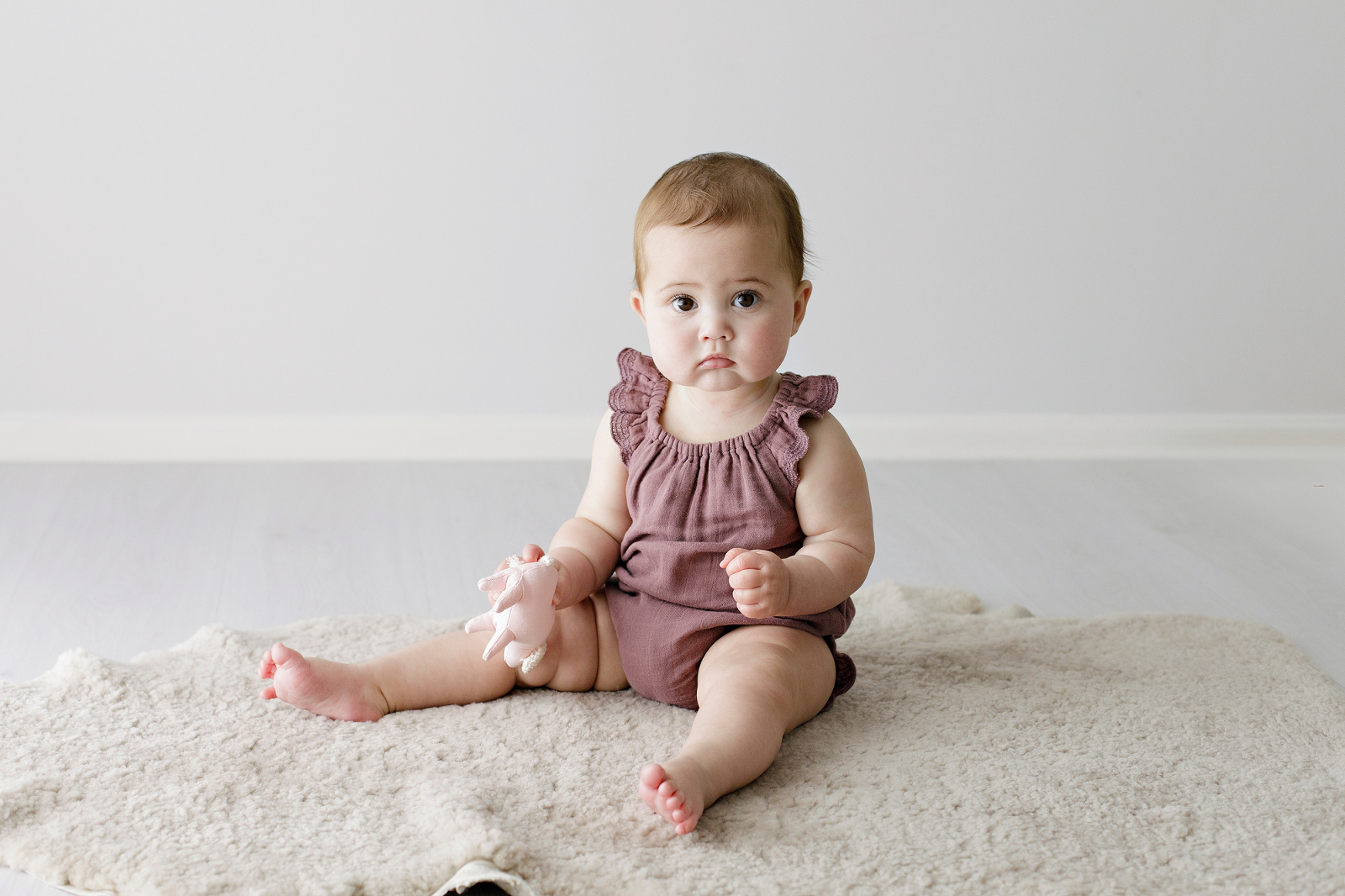 baby girl in a plum coloured romper holding a little pig rattle